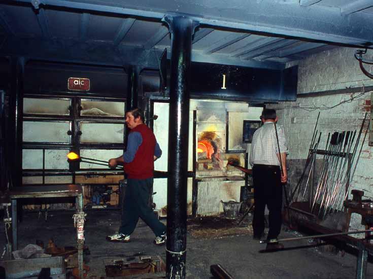 Fig 1. A modern glassblowers workshop. The worker on the left is taking a gather of glass to be blown on the metal mavering slab.