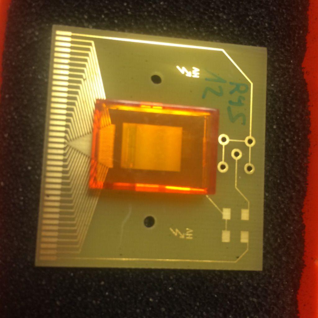 Figure 2: The ROC4SENS in the laboratory. For testing it has an orange cover for protection and sits atop a carrier board. The 35 wire bonds connect the readout chip to the carrier board.