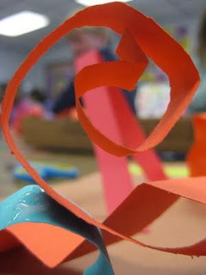 The full sheet of construction paper will act as their base, while the strips will create the sculpture. Take one strip of paper and fold both ends to create feet for the paper to stand on.