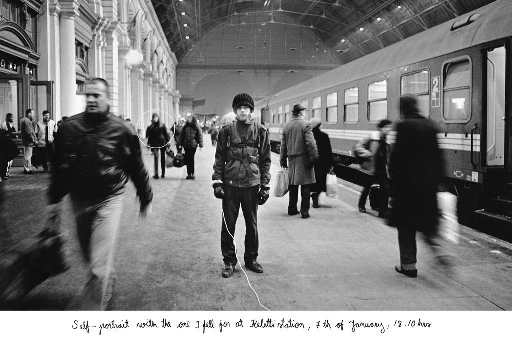 There are people walking in the background, the picture takes place in a train station and there is a train on the left side of the person. The photograph is more of a snap -shot.