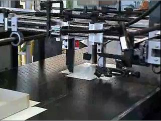 The wraps, adhesive-side up, move along a conveyor from the left. The lid or base, having been formed in the quad stayer, moves toward the operator from the top of the diagram.