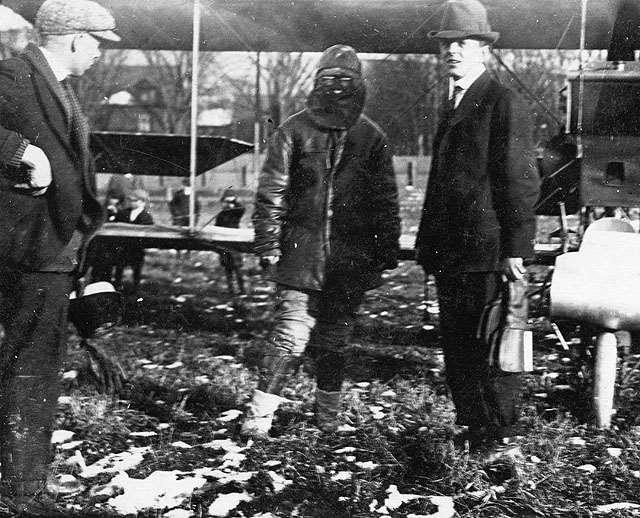 Pilot Ruth Law (center, in heavy flying clothing with full-face mask) poses in front of her Curtiss pusher biplane on the ground at Hornell, New York, November 19, 1916, during her record-breaking